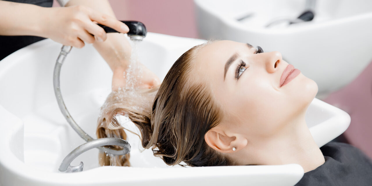Concept beauty salon. Hairdresser washes hair of beautiful blonde girl under tap in wash.