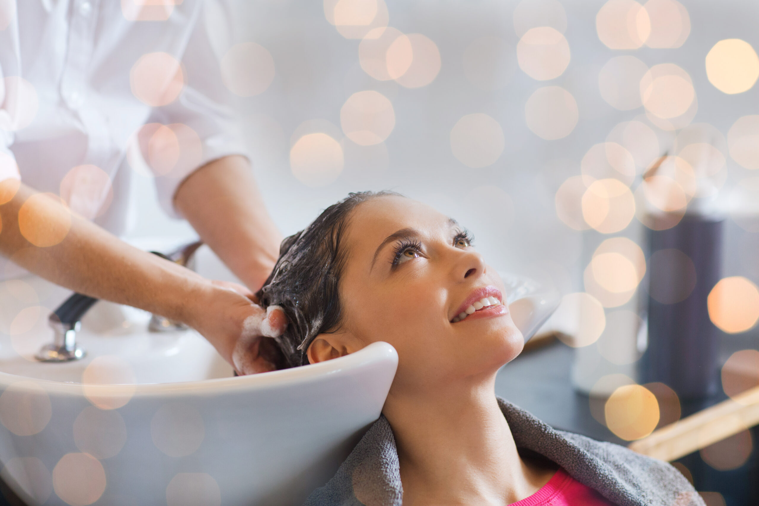 beauty, hair care and people concept - happy young woman with hairdresser washing head at hair salon over holidays lights